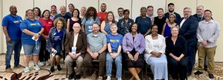 The newly-elected 2025-2027 Executive Council is photographed, with five people seated in the front and the rest standing behind them. They are in front of a beige wall with a tan carpet featuring a circular design with lighter tan and dark brown colors.