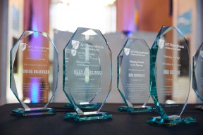 In focus are five of the glass Education Champion Awards, presented to AFTPA members on Saturday, July 12, 2025. Each award is inscribed with the AFTPA logo at the top, the award name in cursive below, the recipient's name in large capital letters in the center, and a description of the award at the bottom. The awards are octagonal, and are set on a table with a black tabletop. The background is blurred.